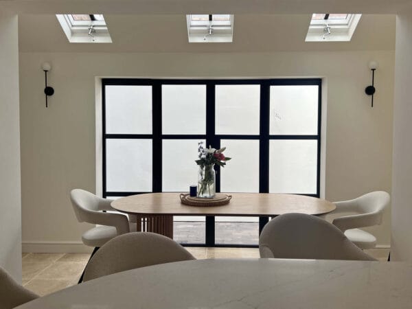 Contemporary dining area with round wooden table, boucle chairs, and black-framed glazing designed by Megan Ace Interiors